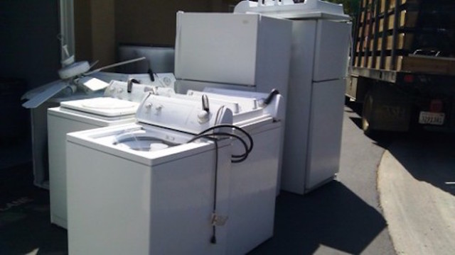 The image shows several old white appliances, including what appears to be washers, dryers, and other household equipment, sitting outside near a truck, likely awaiting disposal or recycling.