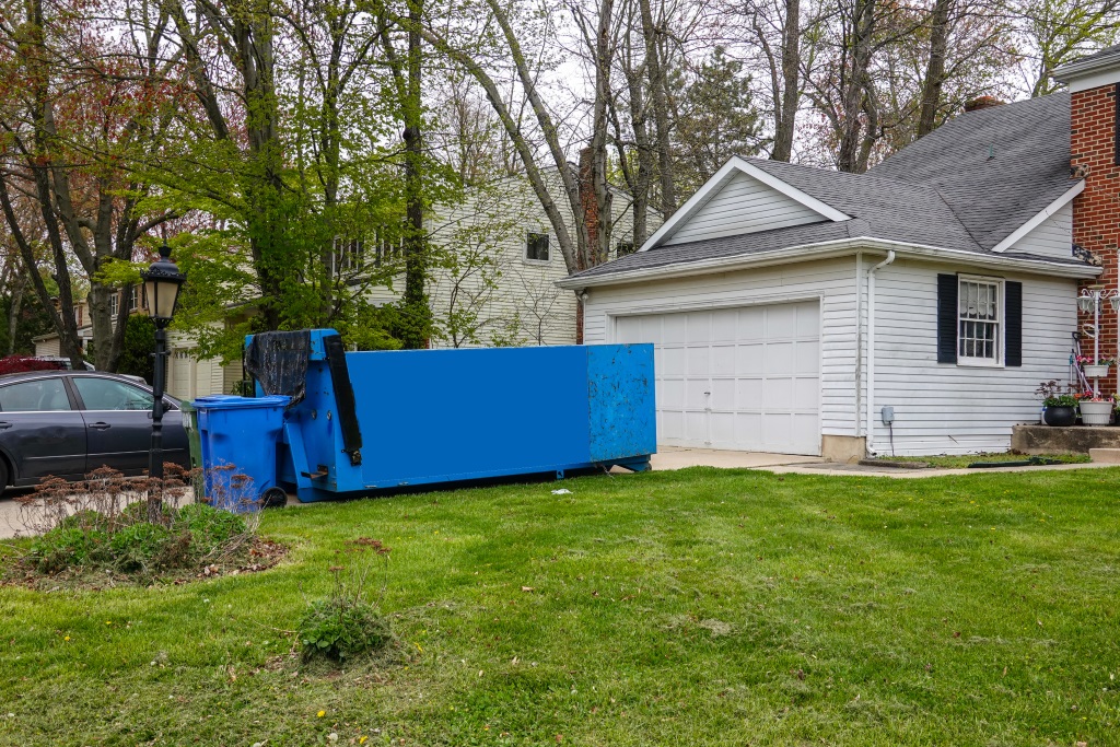 The image shows a bright blue construction dumpster positioned on the green lawn in front of a white residential house with a garage, surrounded by trees in what appears to be a suburban neighborhood.