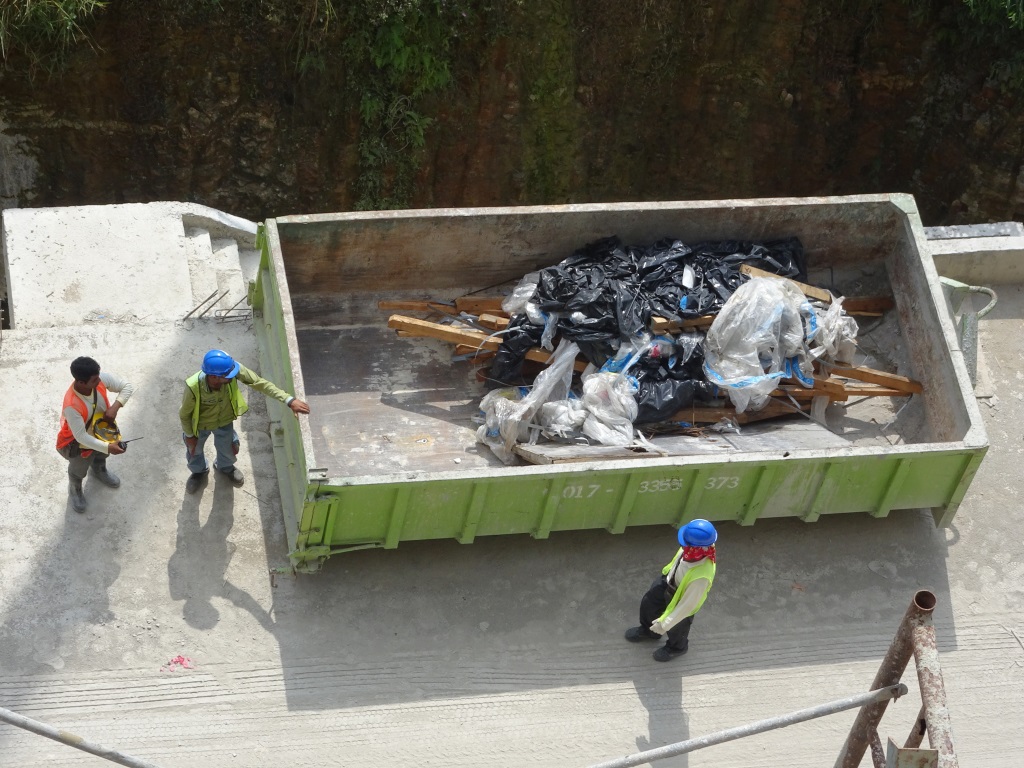 The image shows three construction workers in high-visibility clothing standing around a large green dumpster filled with construction debris including trash bags and wooden materials on what appears to be a construction site.